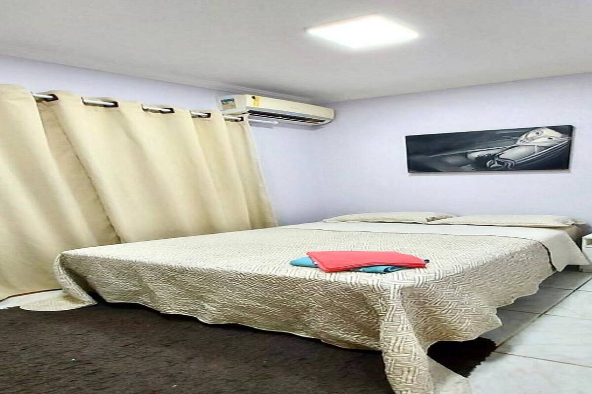 Een slaapkamer met een bed, gordijnen, een airconditioningapparaat, een nachtkastje en een schilderij aan de muur.