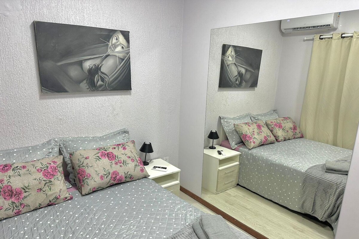 Une chambre avec deux lits, des coussins à motifs floraux, une table de chevet avec une lampe, un miroir et un climatiseur fixé au mur.