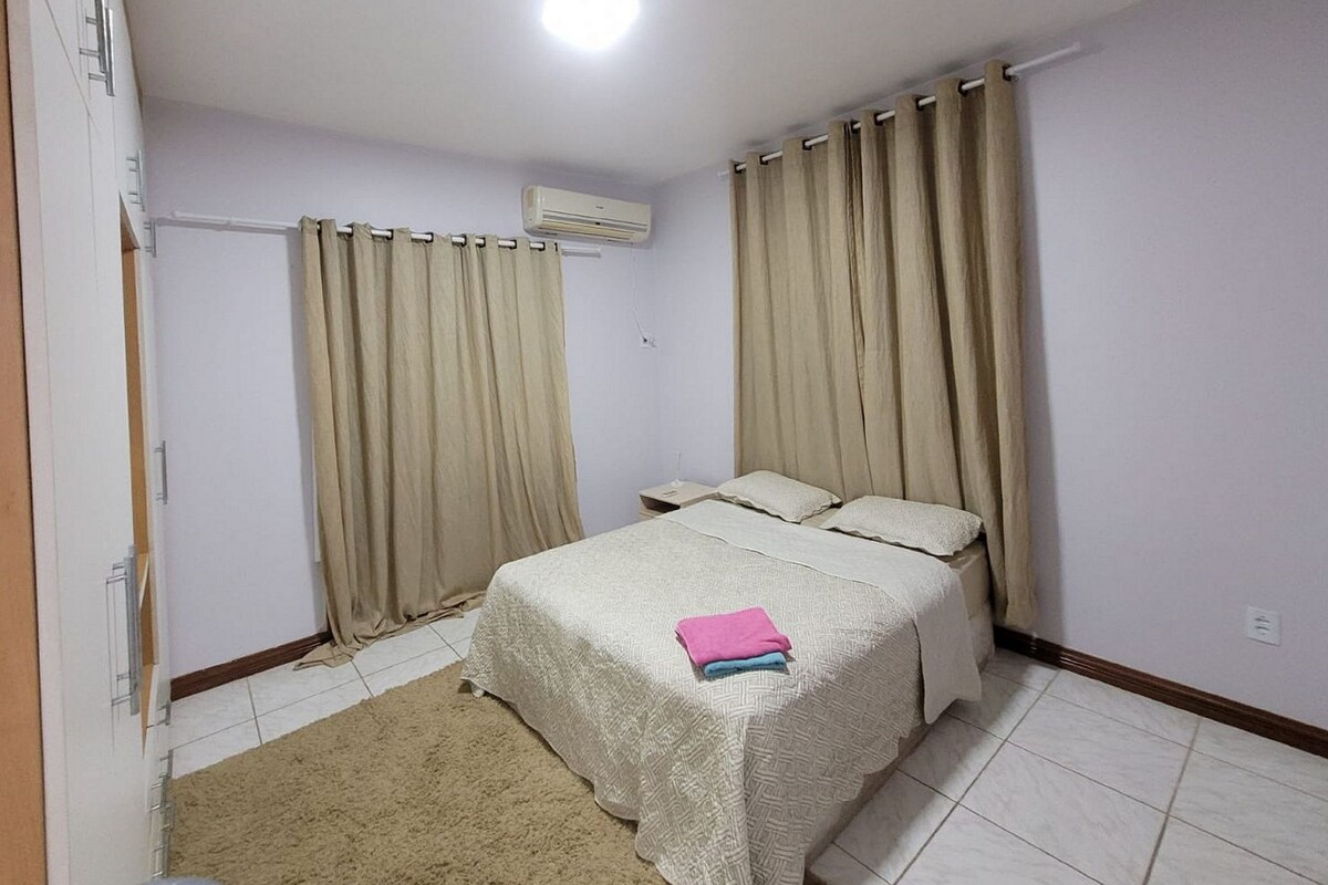 Une chambre à coucher avec un lit, des rideaux beiges, un appareil de climatisation et un sol carrelé.
