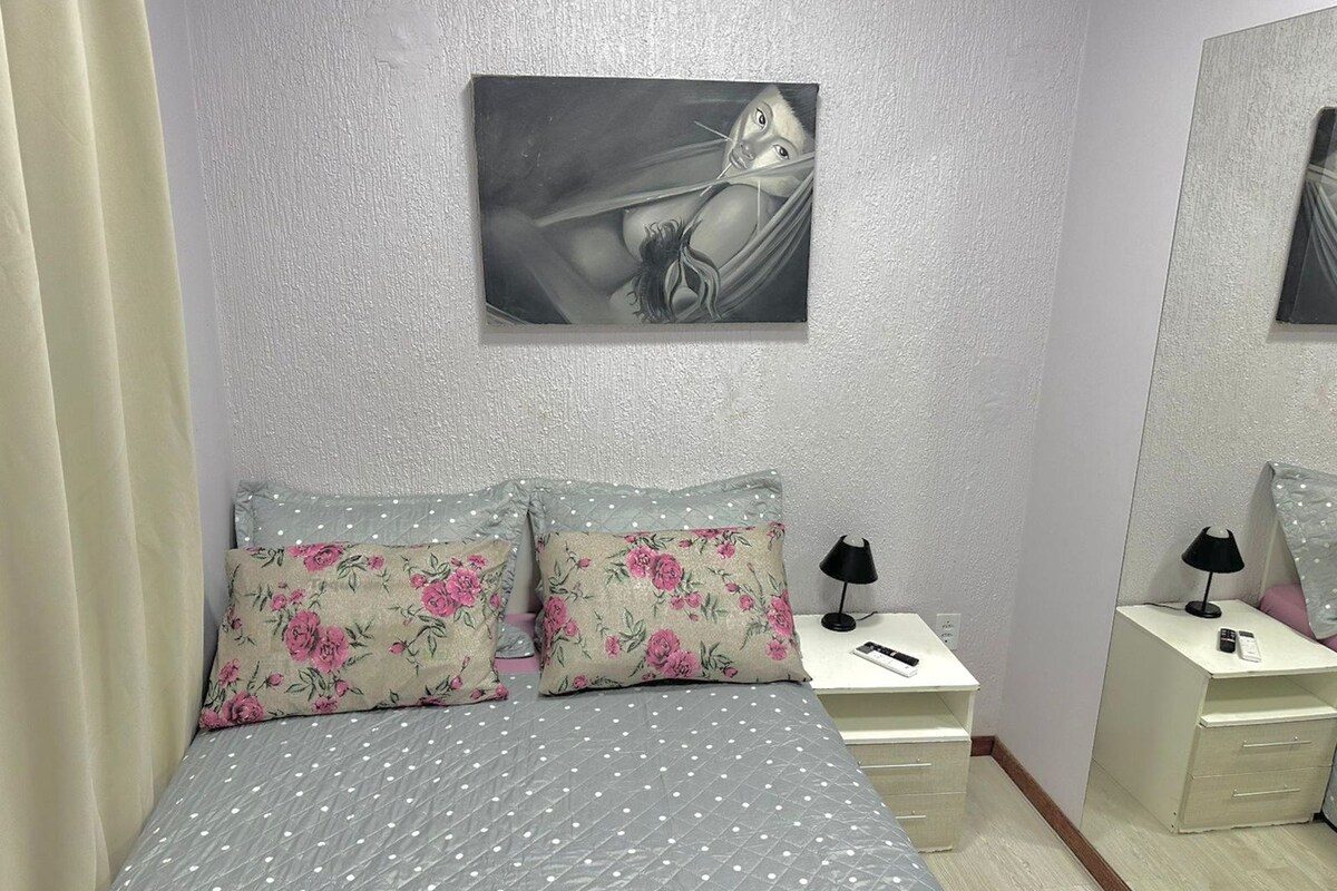 Een slaapkamer met een bed, twee kussens, een nachtkastje, een lamp, een spiegel en een schilderij aan de muur.