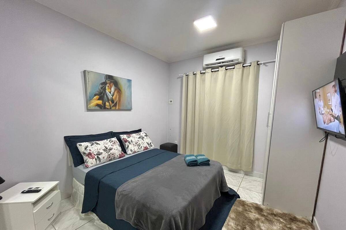 Een slaapkamer met een bed, nachtkastje, televisie, airconditioning en een schilderij aan de muur.