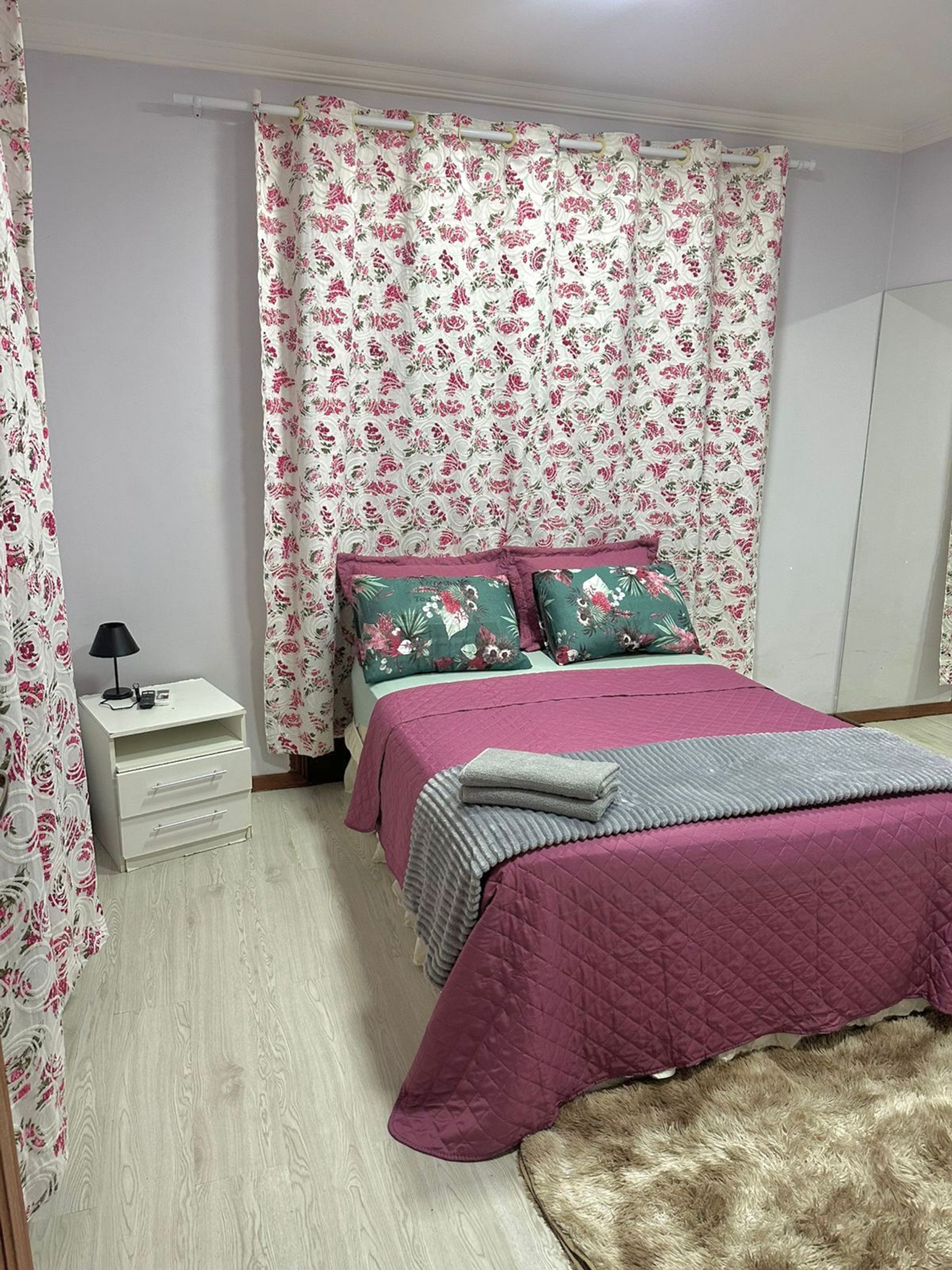 Un dormitorio con una cama, una mesita de noche, una lámpara y cortinas con motivos florales.