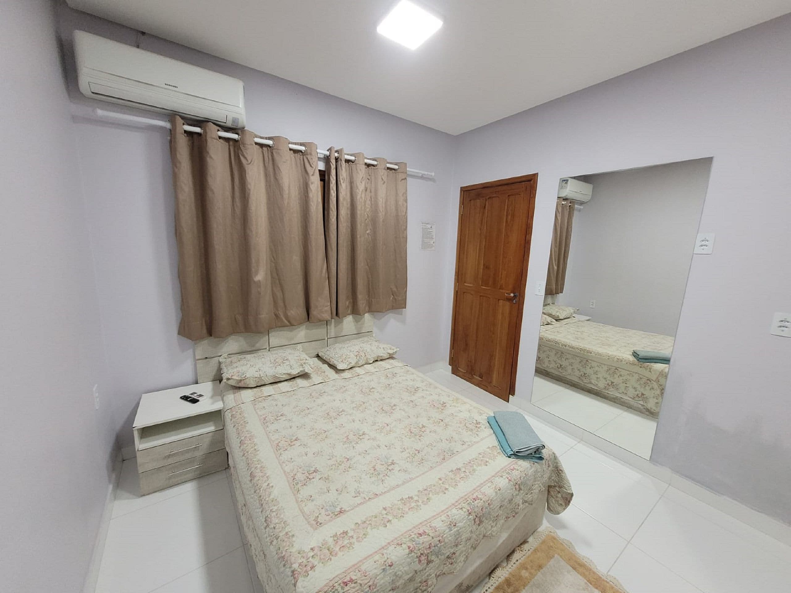 Un dormitorio con una cama, una mesita de noche, un equipo de aire acondicionado, una puerta de madera y un espejo que refleja la cama.