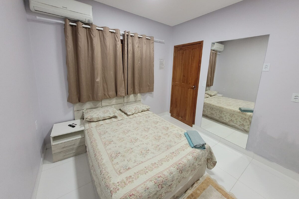 Quarto com cama, mesa de cabeceira, aparelho de ar condicionado, porta de madeira e espelho refletindo a cama.