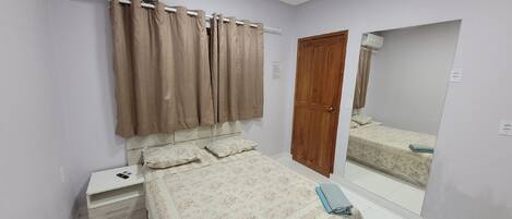 Un dormitorio con una cama, una mesita de noche, un equipo de aire acondicionado, una puerta de madera y un espejo que refleja la cama.