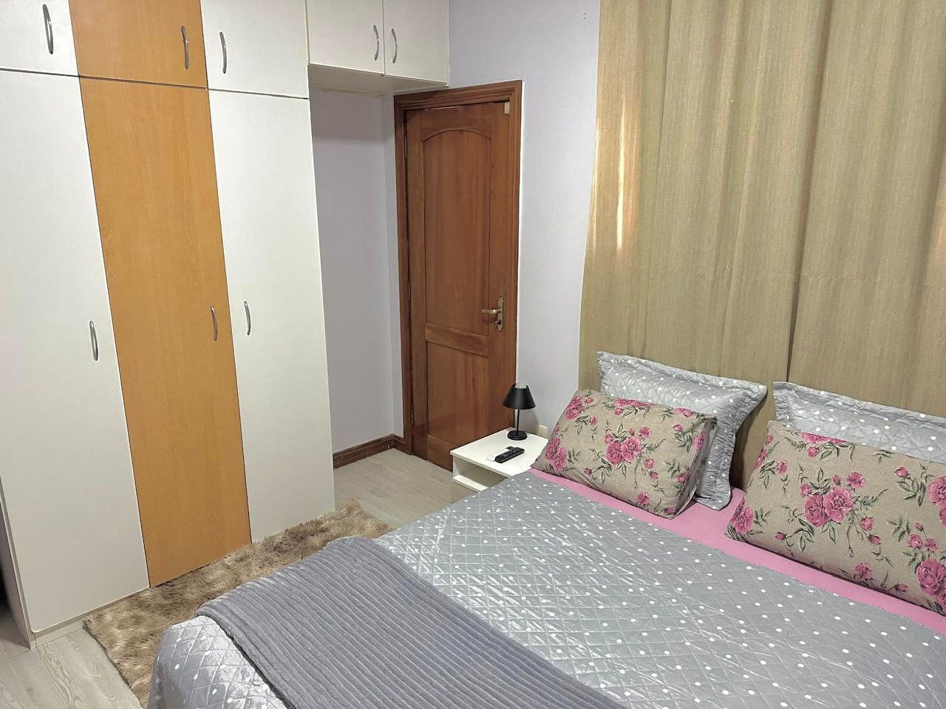 A bedroom with a bed, two pillows, a nightstand, a lamp, a wardrobe, and a door.