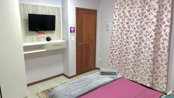 Un dormitorio con cama, televisor montado en la pared, puerta de madera y cortina con motivos florales.