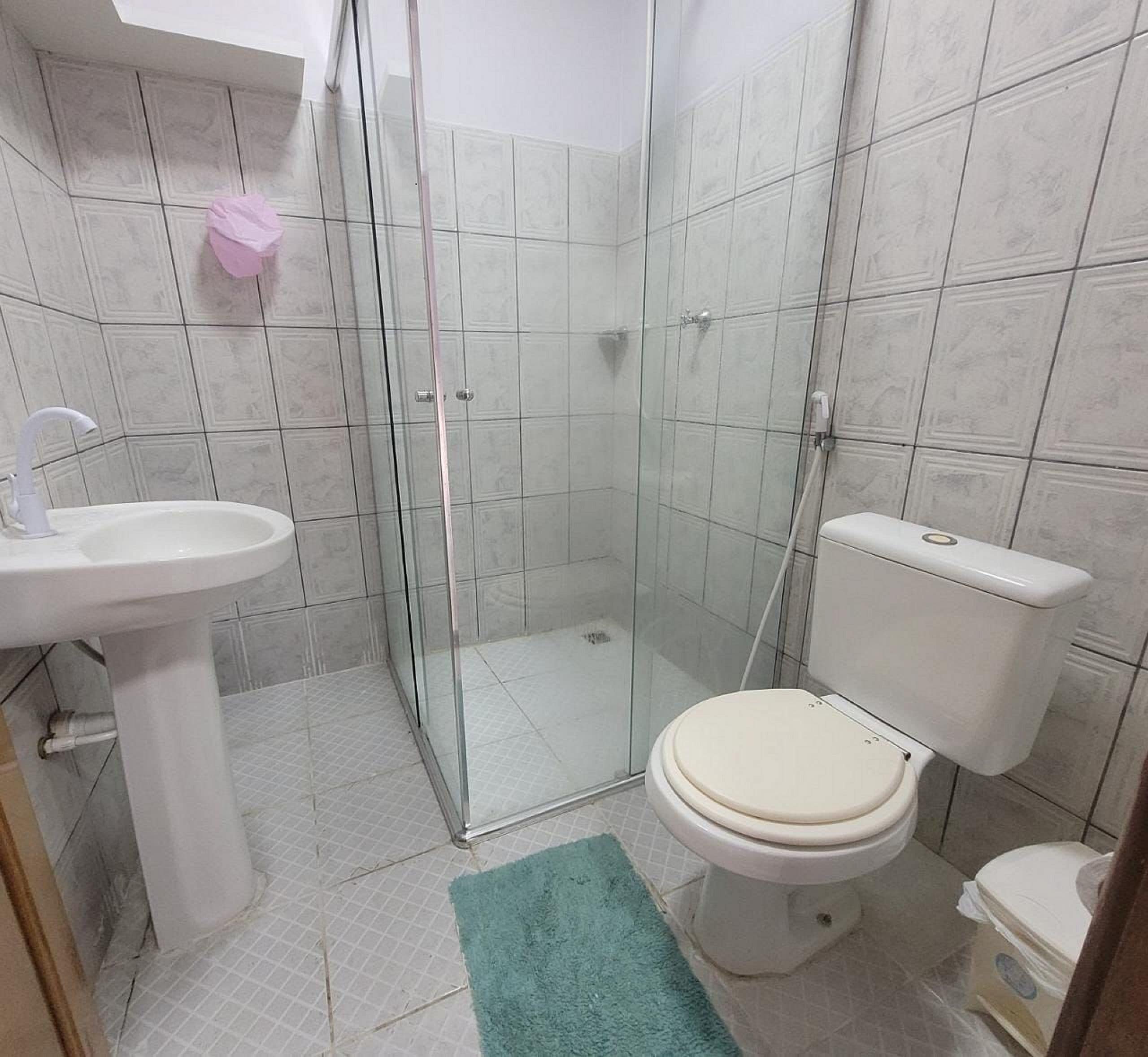 A bathroom with a glass shower enclosure, a white sink, a toilet, and a green bath mat.