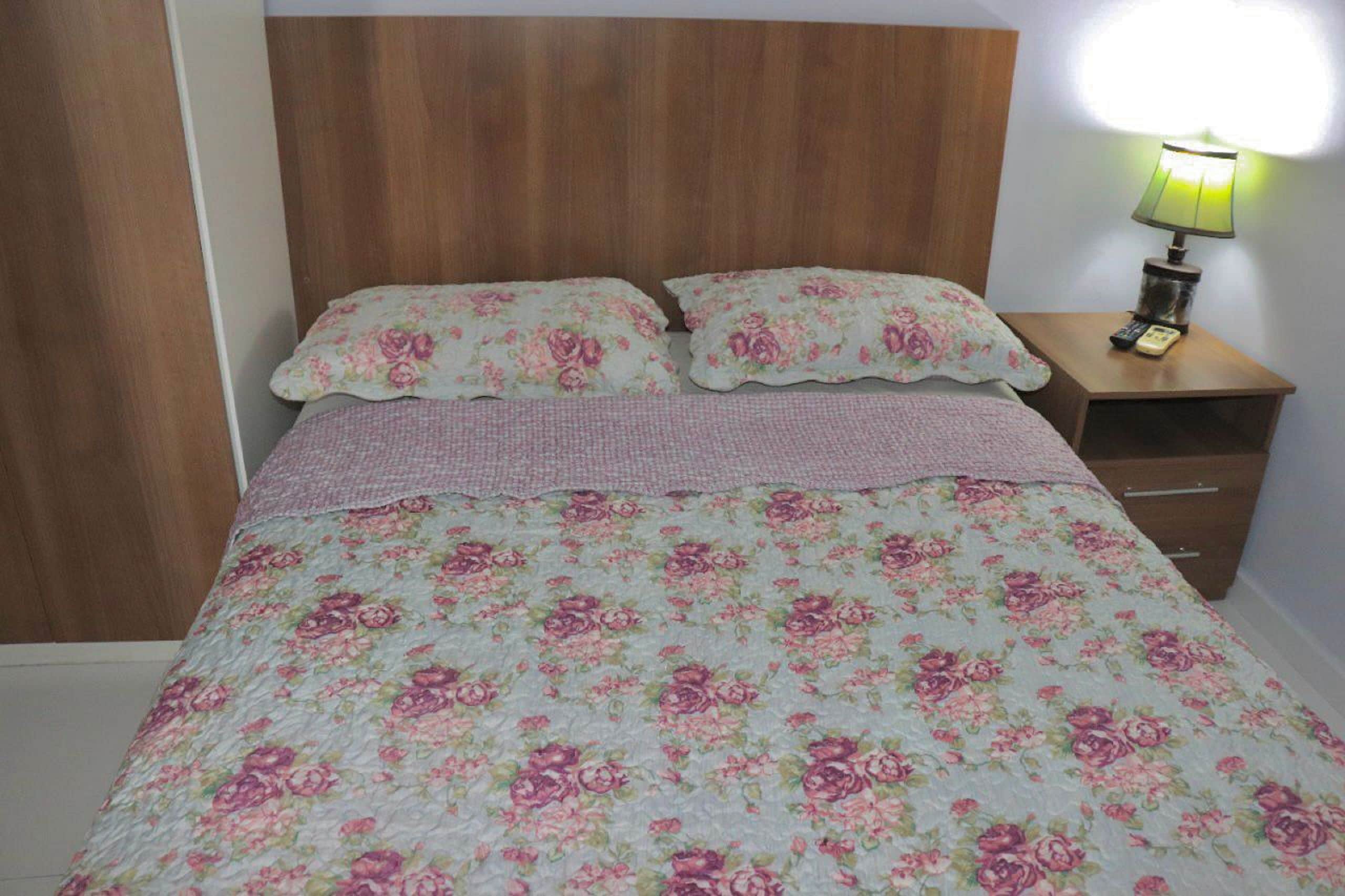 Ein Bett mit floraler Bettwäsche, einem Holzkopfstück und einem Nachttisch mit Lampe und Fernbedienung.