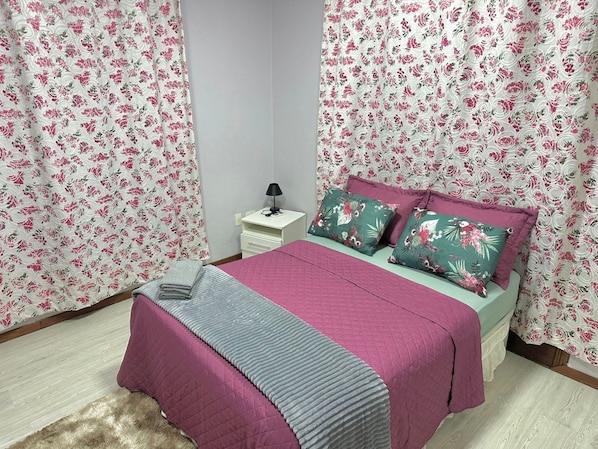 A bedroom with floral curtains, a bed with pink and green bedding, a nightstand with a lamp, and a small drawer.