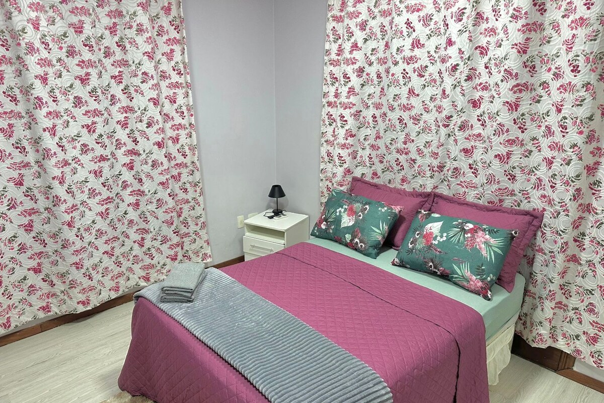 Une chambre à coucher avec des rideaux à motifs floraux, un lit avec une literie rose et verte, une table de chevet avec une lampe et un petit tiroir.