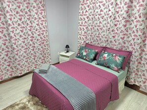 A bedroom with floral curtains, a bed with pink and green bedding, a nightstand with a lamp, and a small drawer.