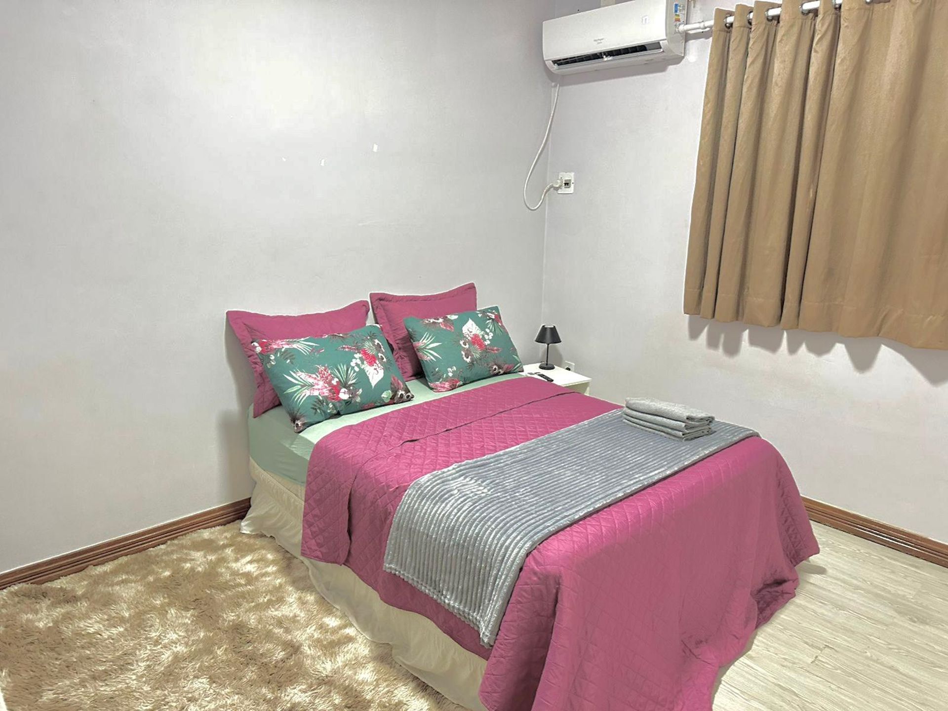 A bedroom with a bed, pillows, a bedside lamp, an air conditioning unit, and a window with curtains.
