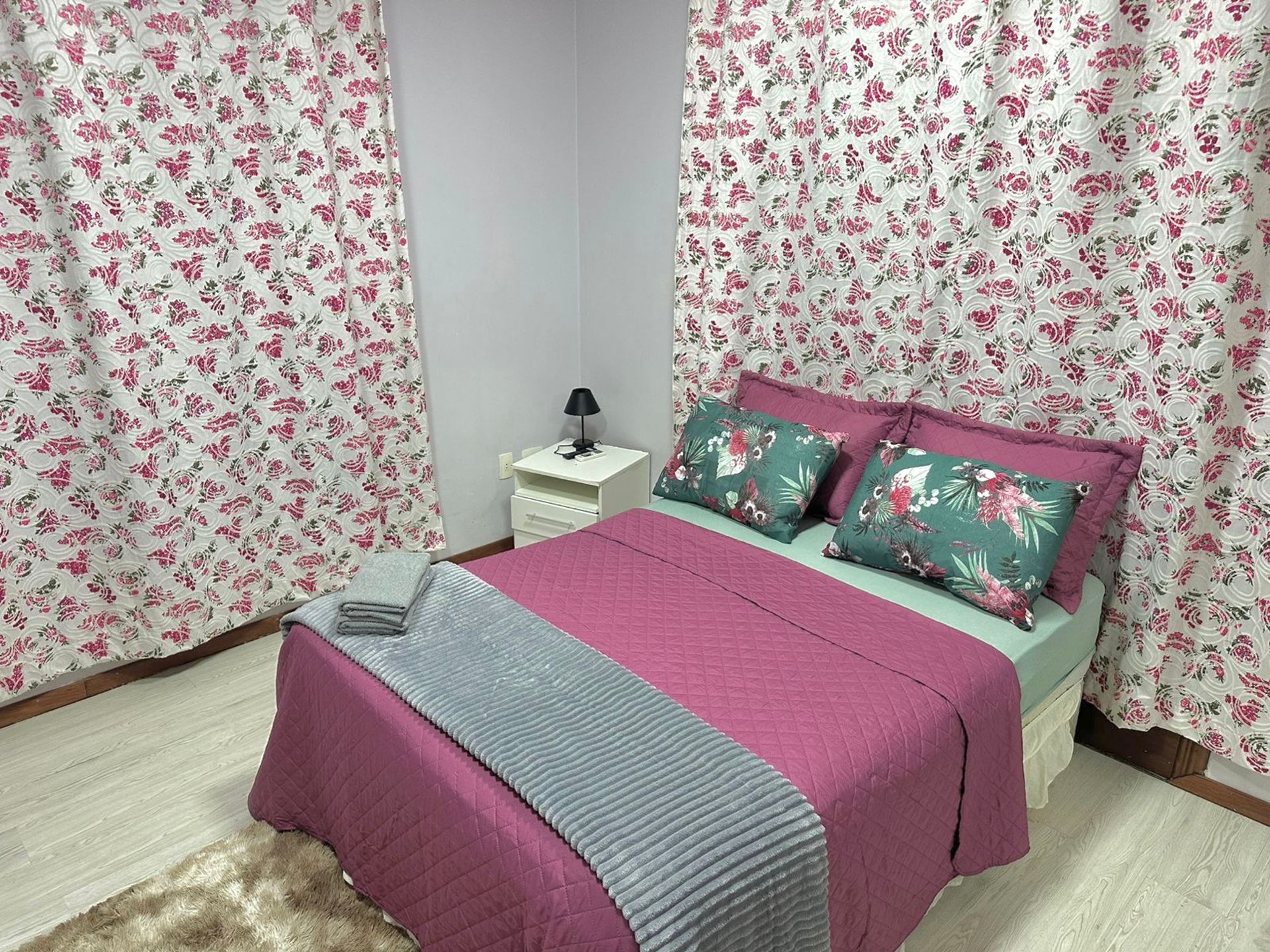 Un dormitorio con cortinas florales, una cama con ropa de cama rosa y verde, una mesita de noche con una lámpara y un cajón pequeño.