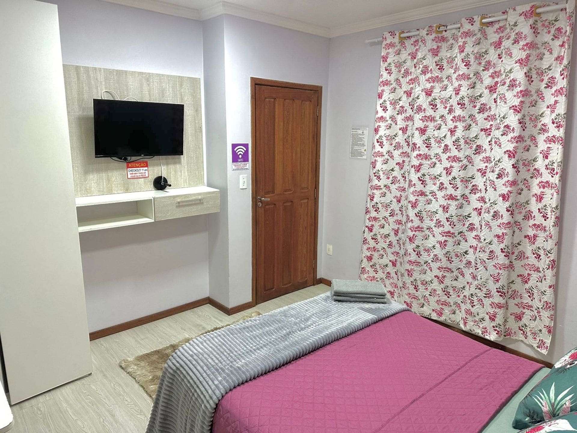 A bedroom with a bed, a TV mounted on a wall, a wooden door, and a floral curtain.