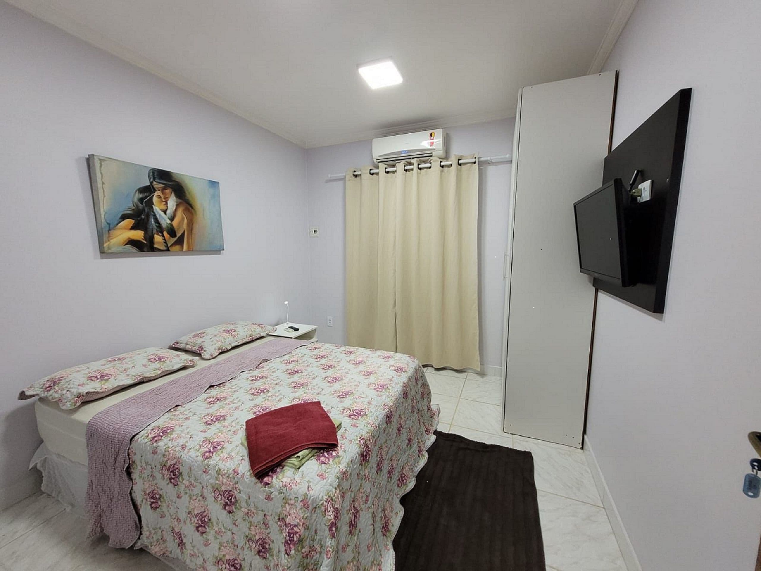 A bedroom with a bed, a painting, an air conditioning unit, a TV, and a wardrobe.