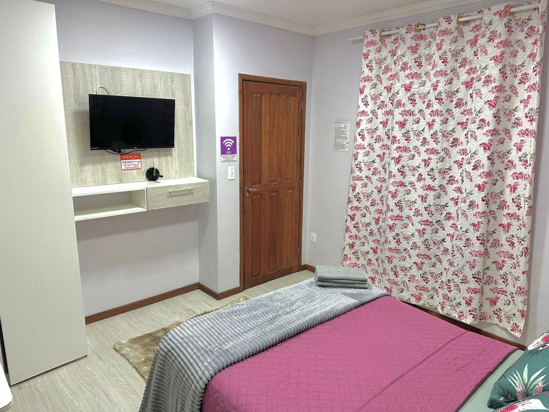 Un dormitorio con cama, televisor montado en la pared, puerta de madera y cortina con motivos florales.
