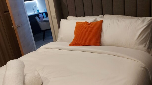 Una cama bien hecha con sábanas blancas y un almohadón naranja, una puerta de madera que da a un baño, y un cabecero moderno.