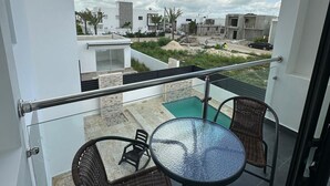 A balcony with two wicker chairs and a glass table, overlooking a swimming pool and residential buildings.