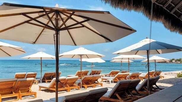 Ein Strandloungebereich mit Holzliegen und großen Sonnenschirmen mit Blick aufs Meer.