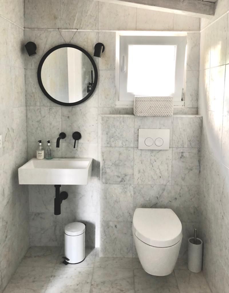 Une salle de bain avec un lavabo blanc, un robinet noir, un miroir rond, des toilettes et une poubelle.