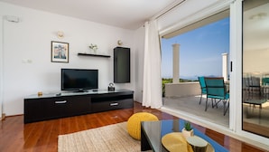 A modern living room with a flat-screen TV, dark wooden flooring, a large window with a view of the outdoors, and a balcony with outdoor furniture. - Apartment Doma - Two Bedroom Apartment with Terrace (Cavtat)