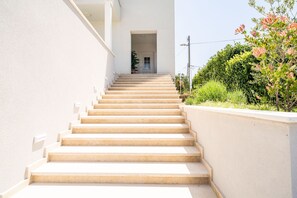 Un escalier beige mène à une porte blanche, qui est entourée de verdure et bordée par un mur blanc.