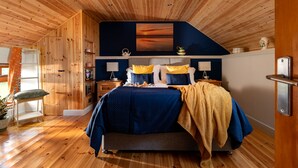 A bedroom with a wooden ceiling, a bed with blue and yellow bedding, a nightstand with a lamp, and a painting above the bed. - Former Coach House in quiet hamlet in Pembrokeshire (Neyland)