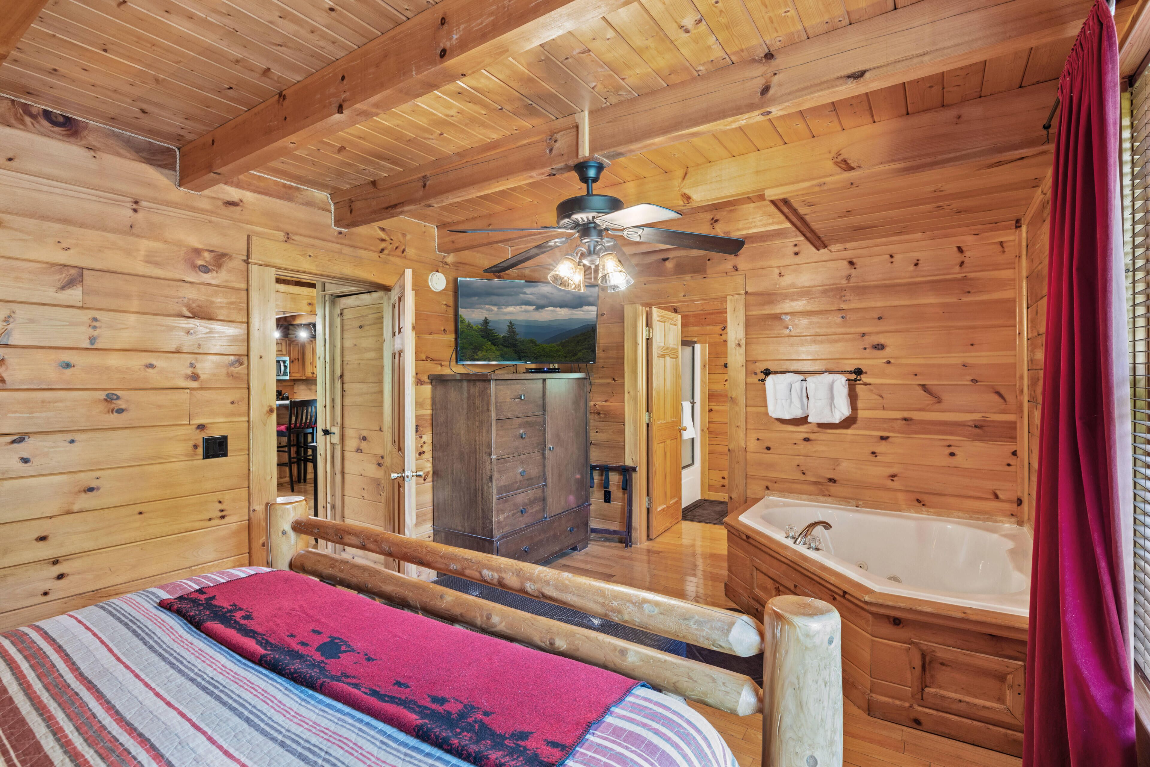 A wooden cabin bedroom with a bed, a ceiling fan, a TV, a wooden cabinet, and a bathroom with a bathtub.