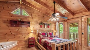 Interior de una cabaña de madera con cama, bañera de madera, ventilador de techo y ventana con vistas a follaje.
