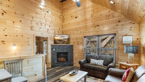 A cozy log cabin interior with a fireplace, wooden furniture, and a view of a mountain through a window.