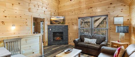 Interior acogedor de una cabaña de troncos con chimenea, muebles de madera y vista a la montaña a través de una ventana.
