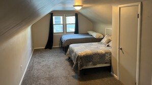 A small, dimly lit attic bedroom with two beds, a window with curtains, and a door. - Walk to Lambeau, Resch, Expo, and Titletown (Green Bay)