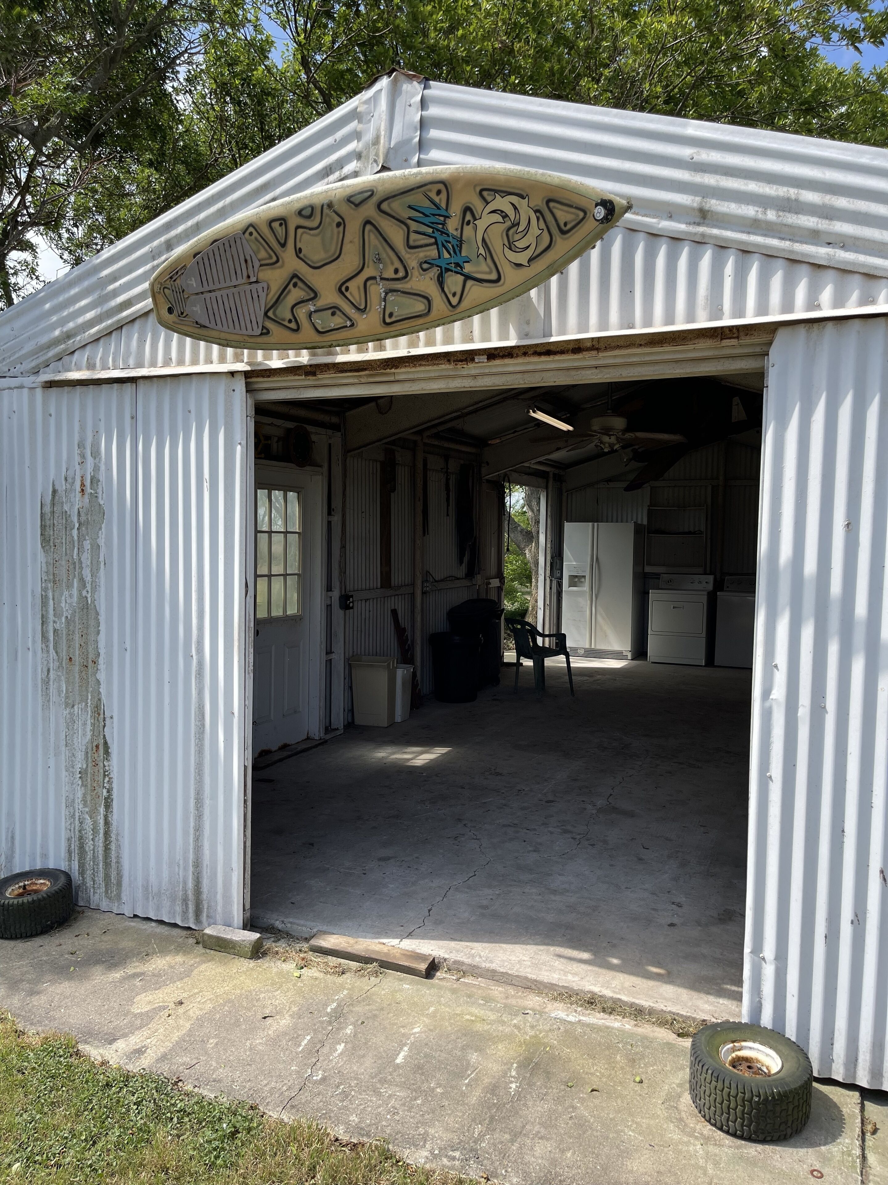 Il s’agit d’un grand hangar ouvert, fait de métal, avec un toit ondulé et une seule porte. À l’intérieur, on trouve divers objets, notamment une chaise, une poubelle et un réfrigérateur.