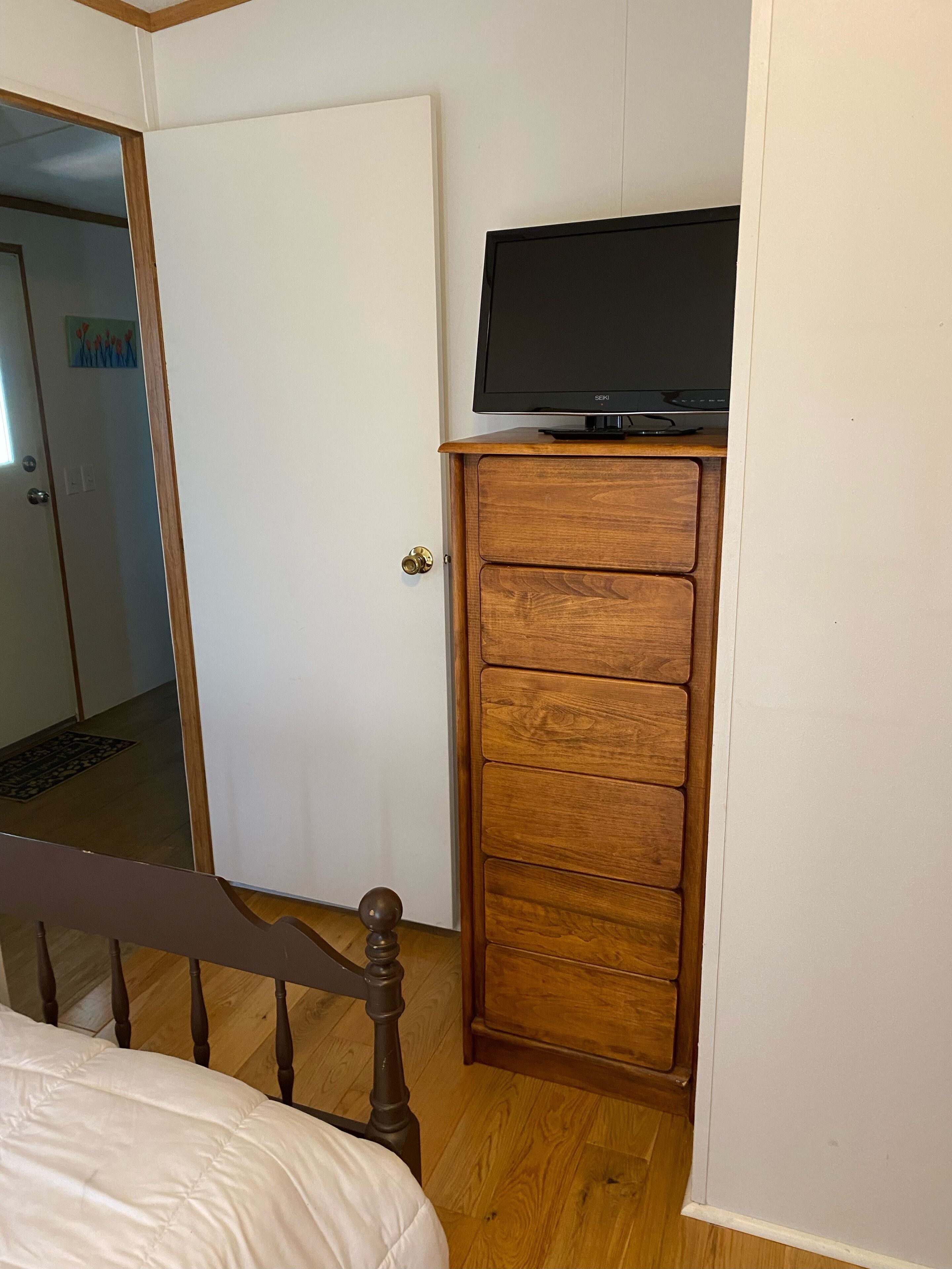 Une chambre avec une armoire en bois, un lit recouvert d’une couette blanche et un téléviseur posé sur le dessus de l’armoire.