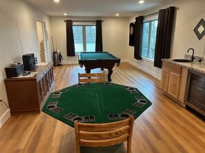 Una mesa de billar en una habitación con piso de madera, una alfombra verde y una zona de cocina con lavaplatos y armarios.
