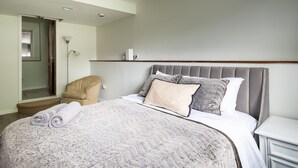 A neatly made bed with a grey headboard, a beige chair, and a bedside table with a lamp.