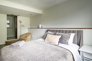 Una cama bien hecha con cabecera gris, una silla color beige y una mesita de noche con lámpara.