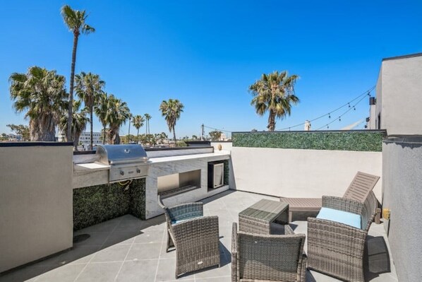 An outdoor patio with a grill, wicker furniture, and palm trees.