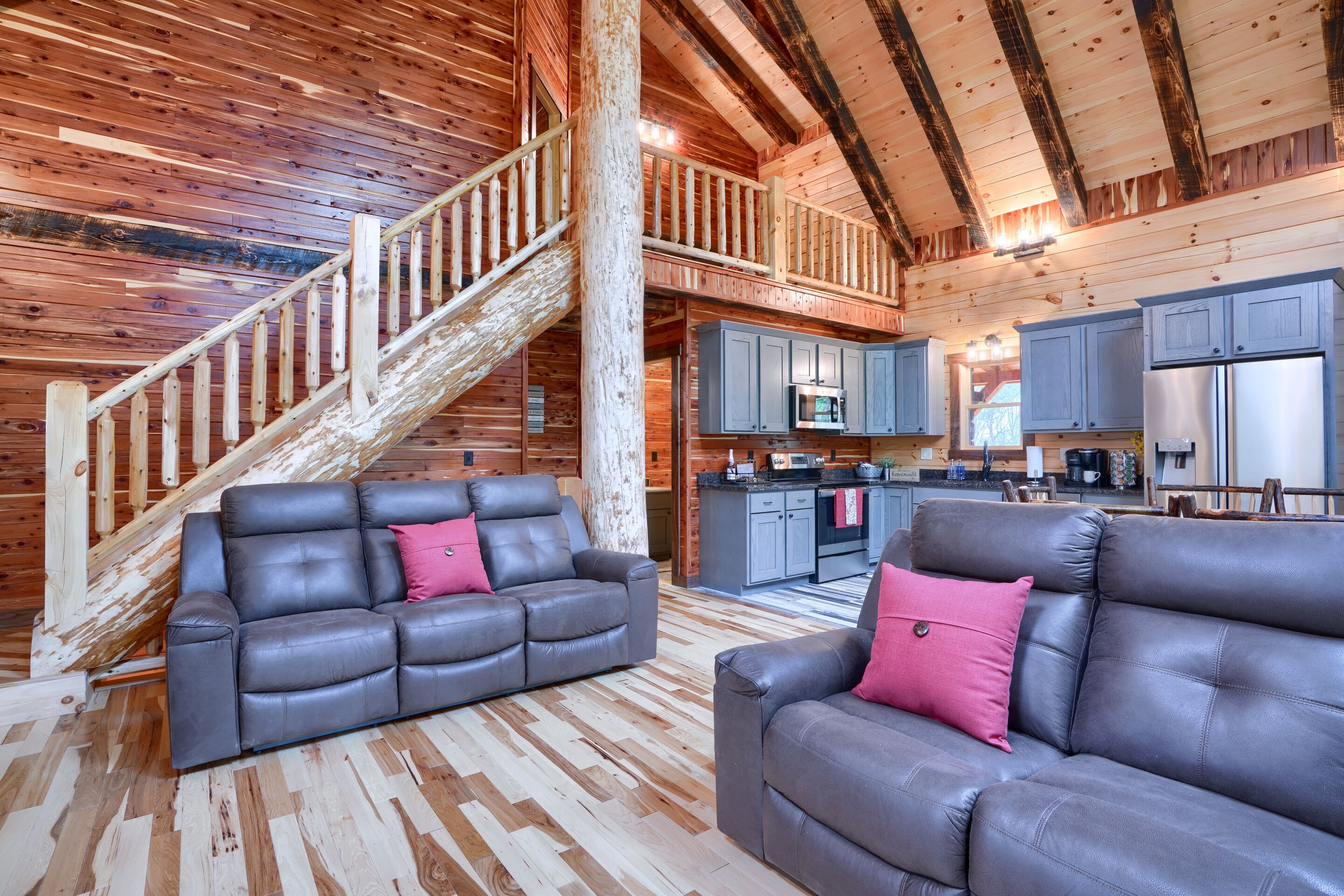 A log cabin interior with a wooden staircase, a kitchen area, and two leather sofas.