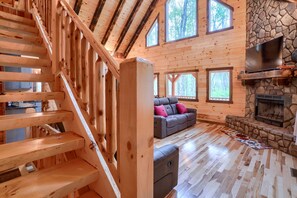 A wooden interior with a staircase, a sofa, a fireplace, and large windows.