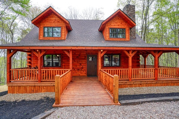 A log cabin with a front porch, two windows, and a stone chimney.