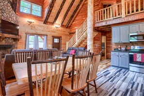 A cabin interior with a stone fireplace, wooden dining table, and a kitchen with modern appliances.