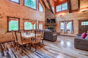 A wooden cabin interior with a dining table, chairs, a fireplace, a sofa, and large windows.