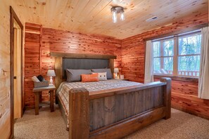 Quarto de cabana de madeira com cama, mesa de cabeceira, abajur e janela com vista para árvores.