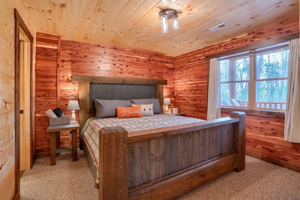 A wooden cabin bedroom with a bed, bedside table, lamp, and a window with a view of trees.