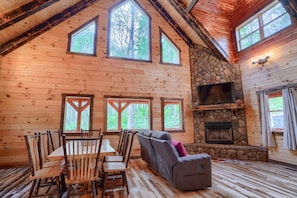 A wooden cabin interior with a dining area, a stone fireplace, and large windows.