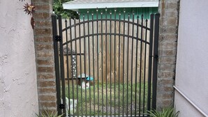 Une entrée fermée par un portail, avec une arche en pierre, une grille en métal et un chemin menant à une construction surmontée d’un toit végétalisé.