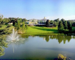 A tranquil lake with a fountain, surrounded by lush greenery and a large house with a visible porch.