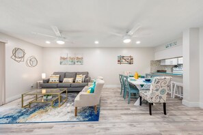 A modern living room with a grey sofa, a glass coffee table, and a dining area with teal chairs and a white table. - Modern~ Ground floor Condo~ 4 mins walk to Beach! Sleeps 6 (Fort Walton Beach)